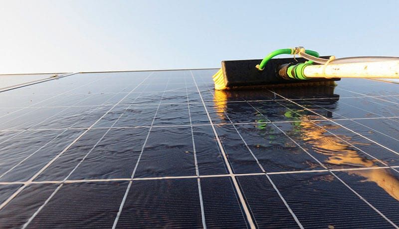 Brush rinsing a solar panel at golden hour, the wet glass reflecting the sky