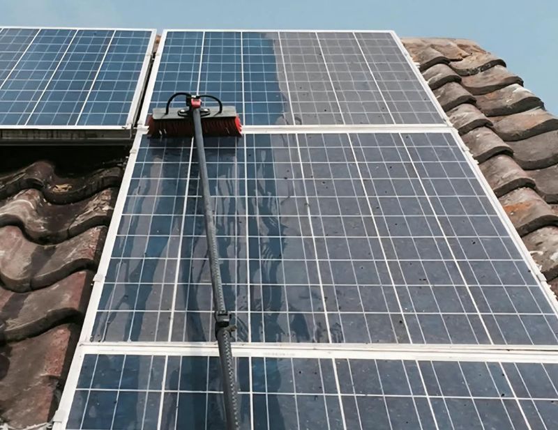 A water-fed brush cleaning rooftop solar panels on a tiled roof, with the clean section visibly darker and clearer